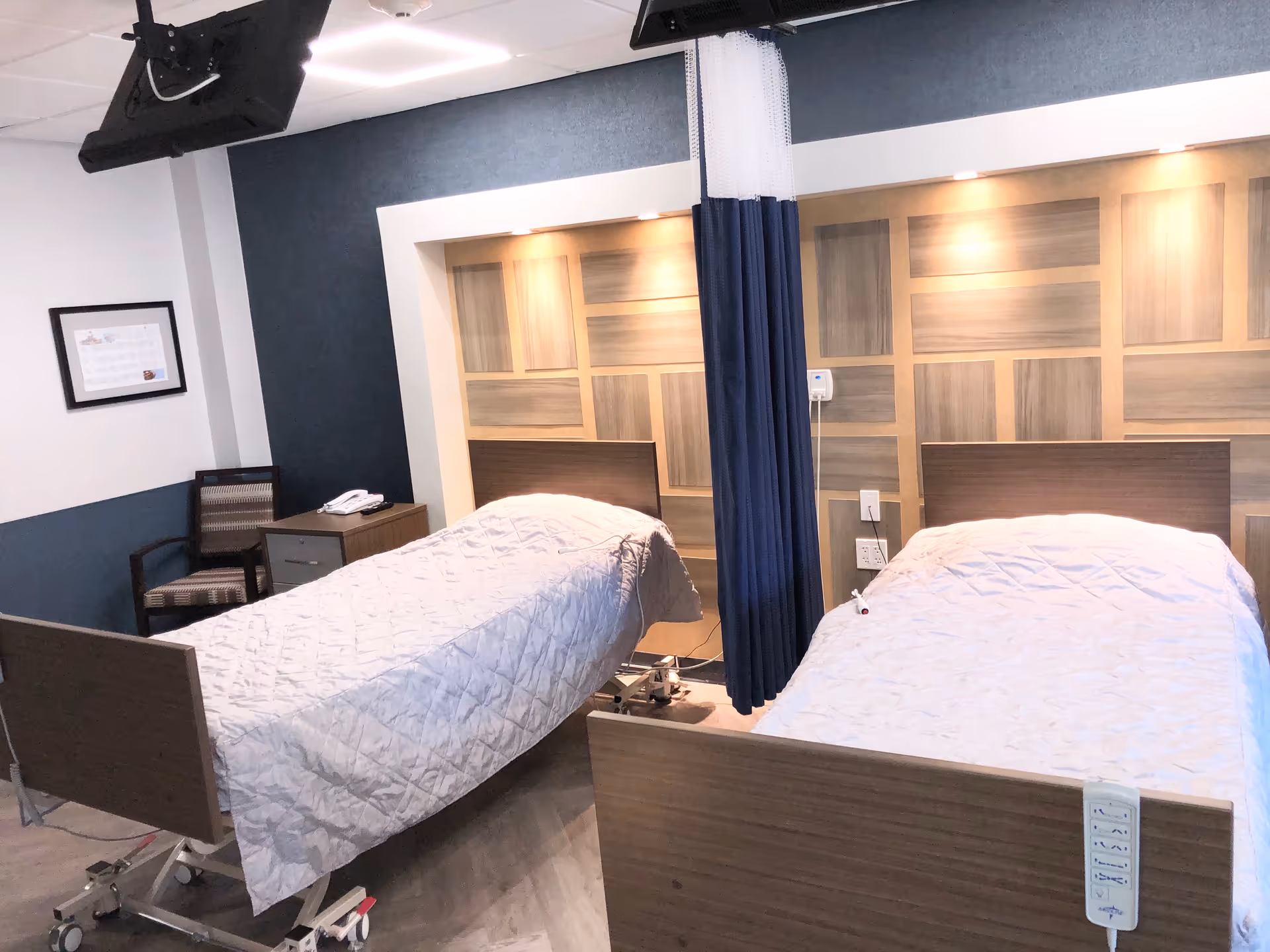 A room with two hospital-style beds covered with white quilts, separated by a blue privacy curtain. The beds have wooden headboards and footboards, and there is a nightstand with a telephone and a chair against the wall. The wall behind the beds features a decorative wood panel design with built-in lighting.