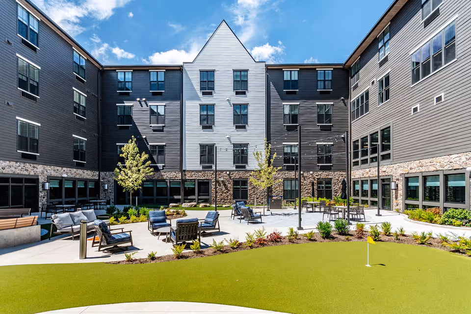 Outdoor courtyard with seating, a small putting green, landscaping and a surrounding multi-story building under a blue sky.