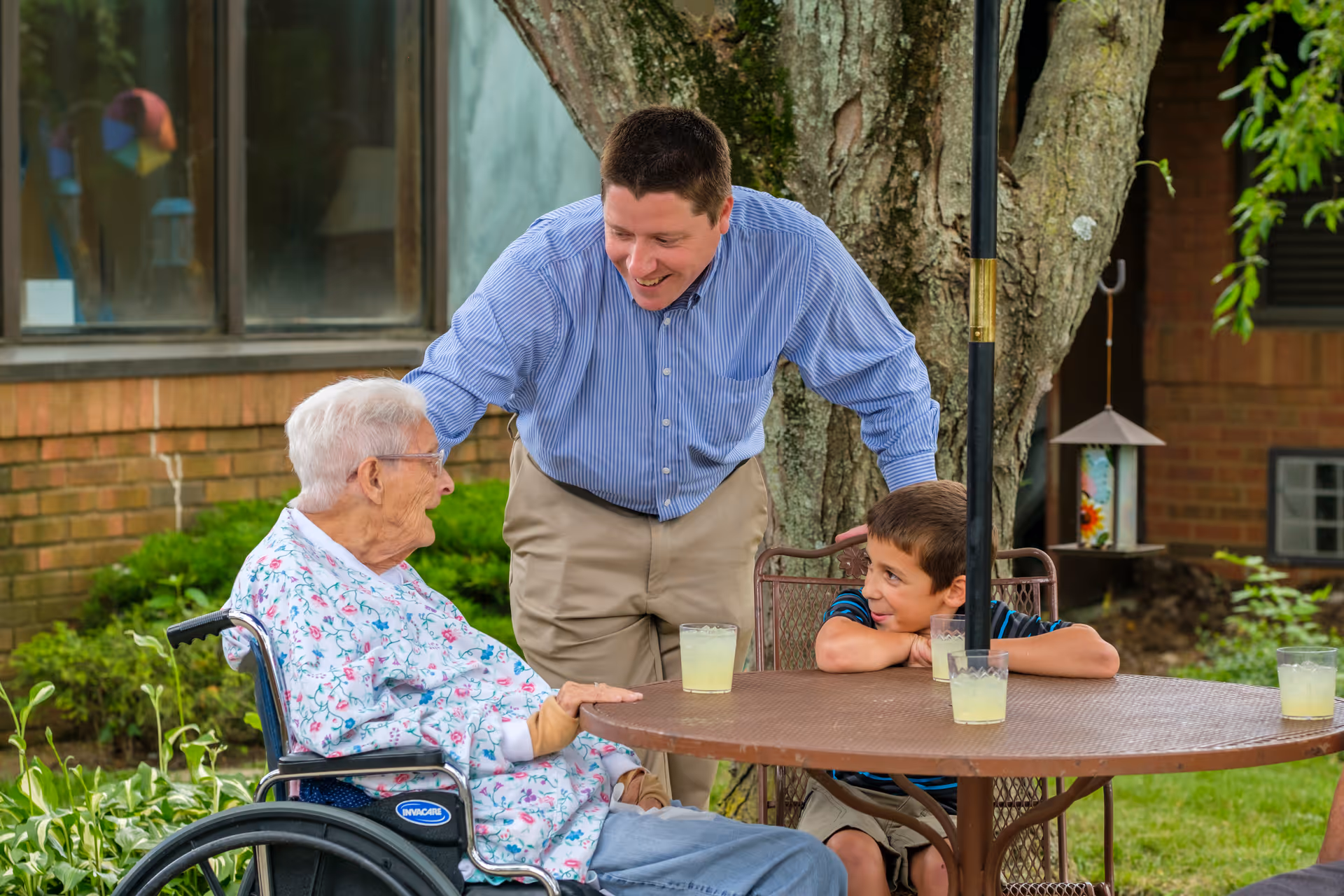 An elderly woman in a wheelchair, a man, and a young boy are gathered around a round outdoor table with glasses of lemonade. They are smiling and engaging in conversation in a garden area with a tree and brick building in the background.