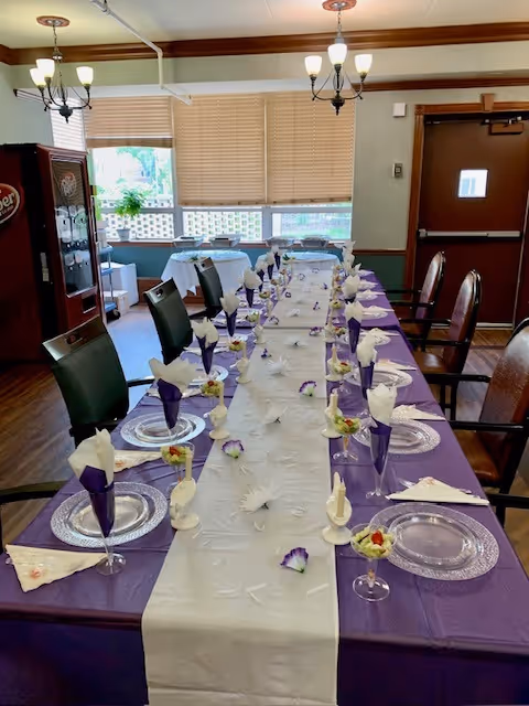 Long communal dining table set for a meal with purple tablecloth, white runner, folded napkins, glass plates and chandeliers in a dining room.