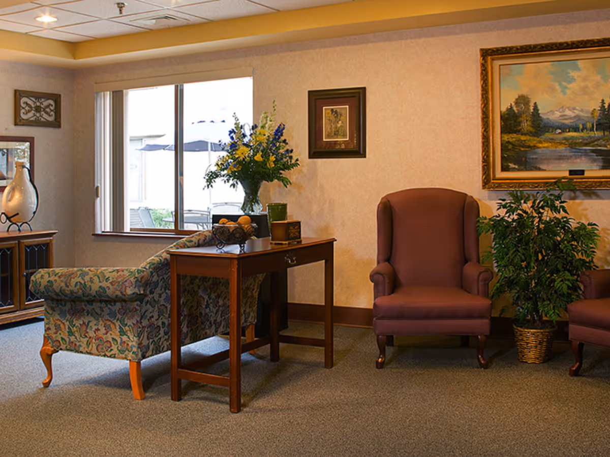 A cozy living room area with a floral patterned sofa, a wooden side table with a vase of flowers and decorative items, two maroon armchairs, a potted plant, and framed artwork on the walls. A window shows an outdoor patio with umbrellas.