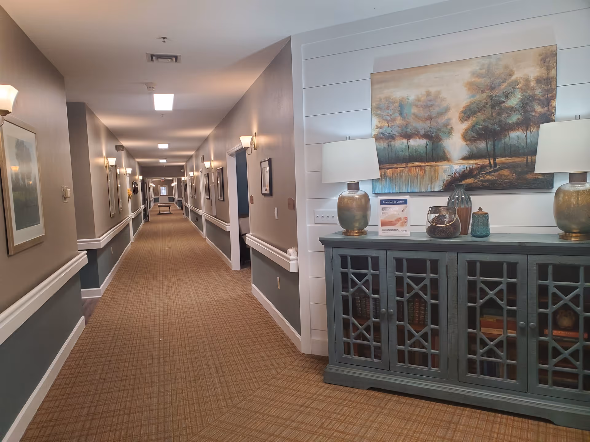 A long, well-lit hallway in a senior living facility with beige and teal walls, carpeted floor, and multiple wall-mounted lights. On the right side near the foreground, there is a blue cabinet with decorative items including two lamps, vases, and a framed landscape painting above it.