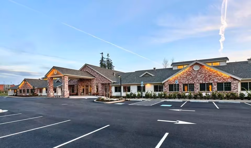 Exterior view of The Rawlin at Riverbend Memory Care facility showing a single-story building with brick and white siding, a covered entrance, and an empty parking lot with marked spaces and directional arrows under a clear sky at dusk.