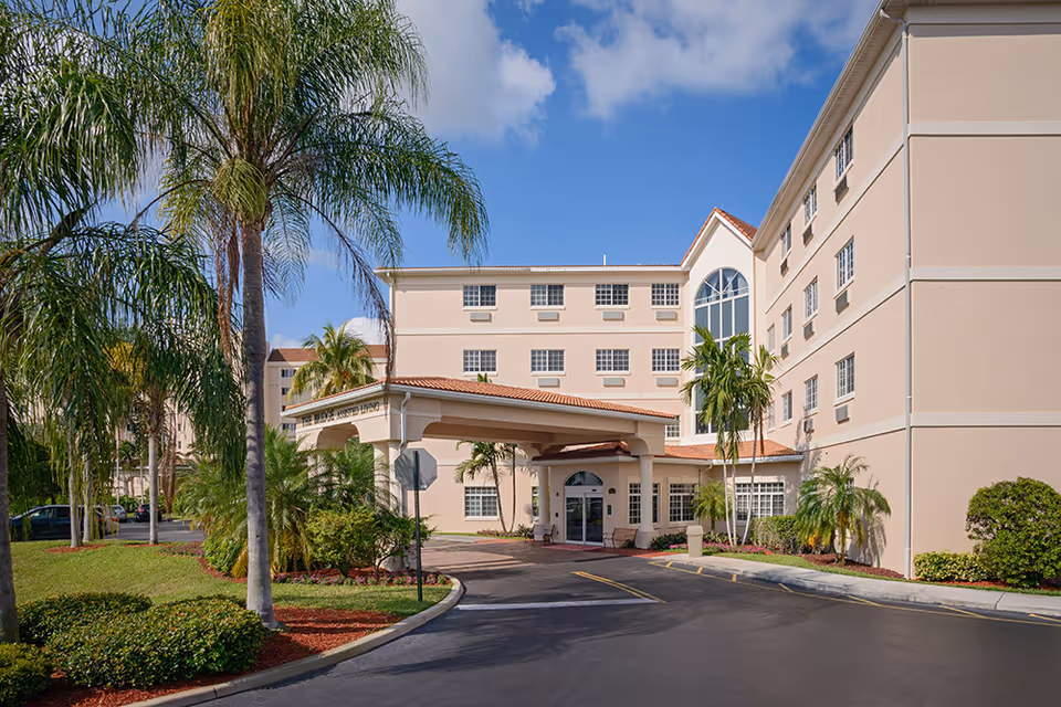 Exterior view of a multi-story senior living facility with beige walls and a red-tiled roof. The entrance has a covered driveway with palm trees and landscaped greenery surrounding the building under a partly cloudy blue sky.