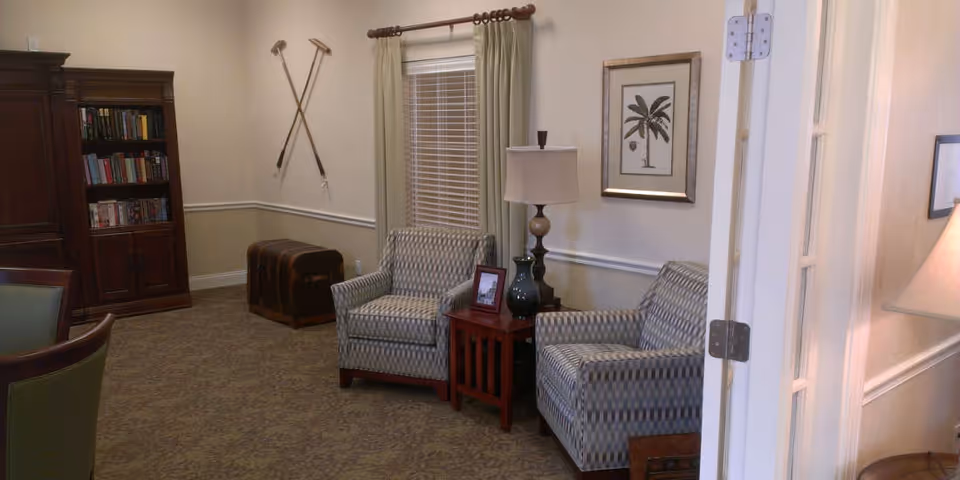 A cozy sitting area in a senior living facility featuring two patterned armchairs separated by a wooden side table with a lamp and decorative items. Behind the chairs is a window with curtains, a framed palm tree picture on the wall, and two crossed walking canes mounted on the adjacent wall. A wooden bookshelf filled with books and a vintage trunk are also visible in the room.