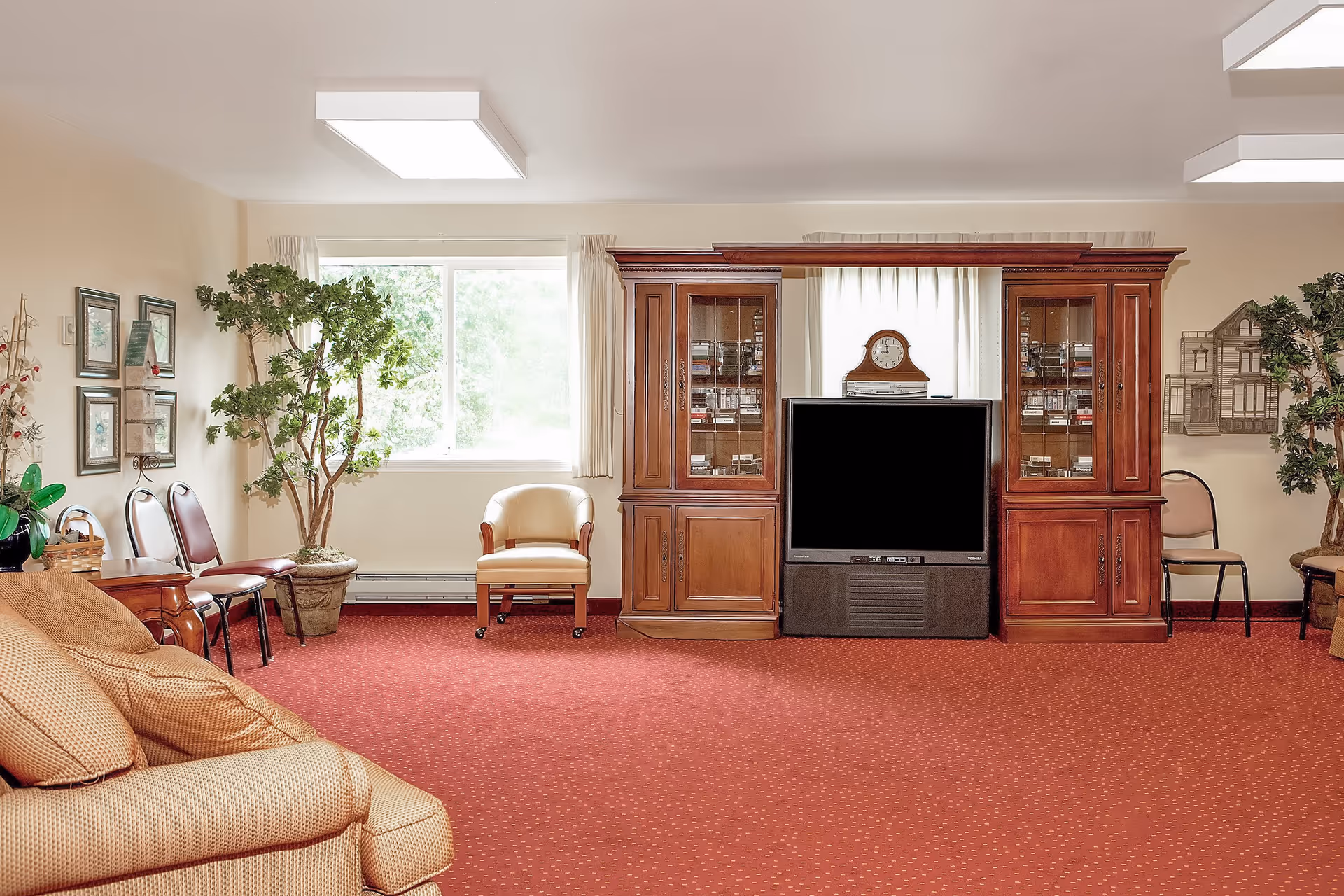 A living room with a large wooden entertainment center holding a television and a clock. The room has a red carpet, beige walls, and a window with light curtains. There are several chairs, a beige sofa, potted plants, and framed pictures on the walls.