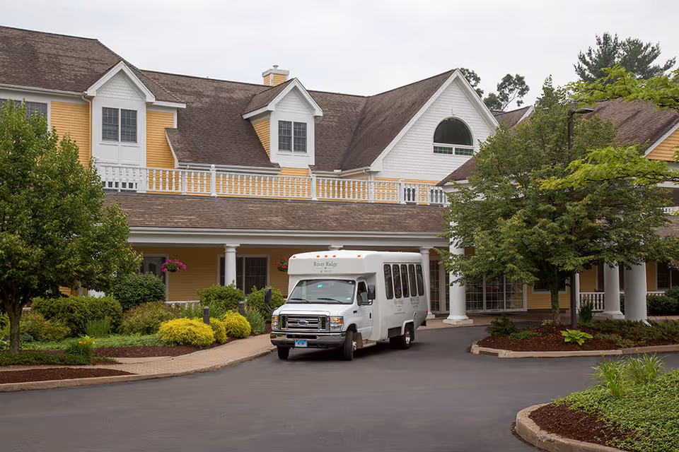 Front entrance of a yellow senior living building with landscaping and a white 'River Ridge' shuttle bus parked under the porte-cochere.