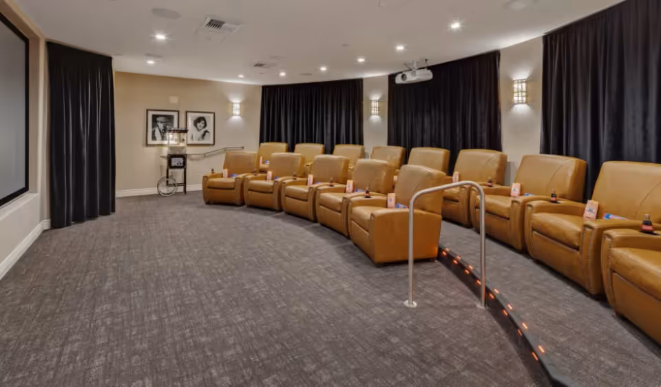 A small theater room with two rows of tan leather recliner chairs facing a large projection screen. The room has dark curtains on the windows, wall sconces for lighting, and a popcorn machine in the back corner. There is a handrail along a step between the two rows of chairs.