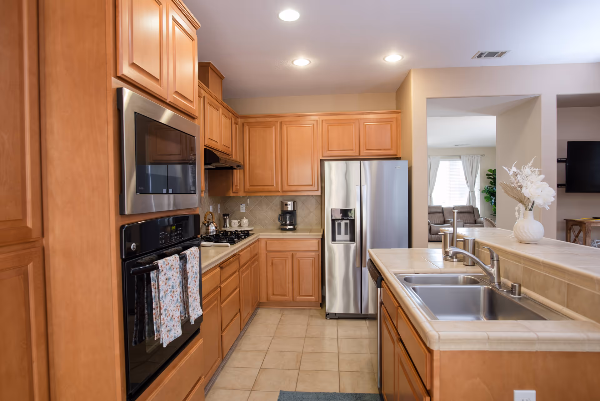 A modern kitchen with wooden cabinets, a stainless steel refrigerator, built-in microwave and oven, a gas stove, and a double sink on a tiled countertop. The kitchen opens into a living area with a sofa and a TV mounted on the wall. There is a vase with white flowers on the counter.