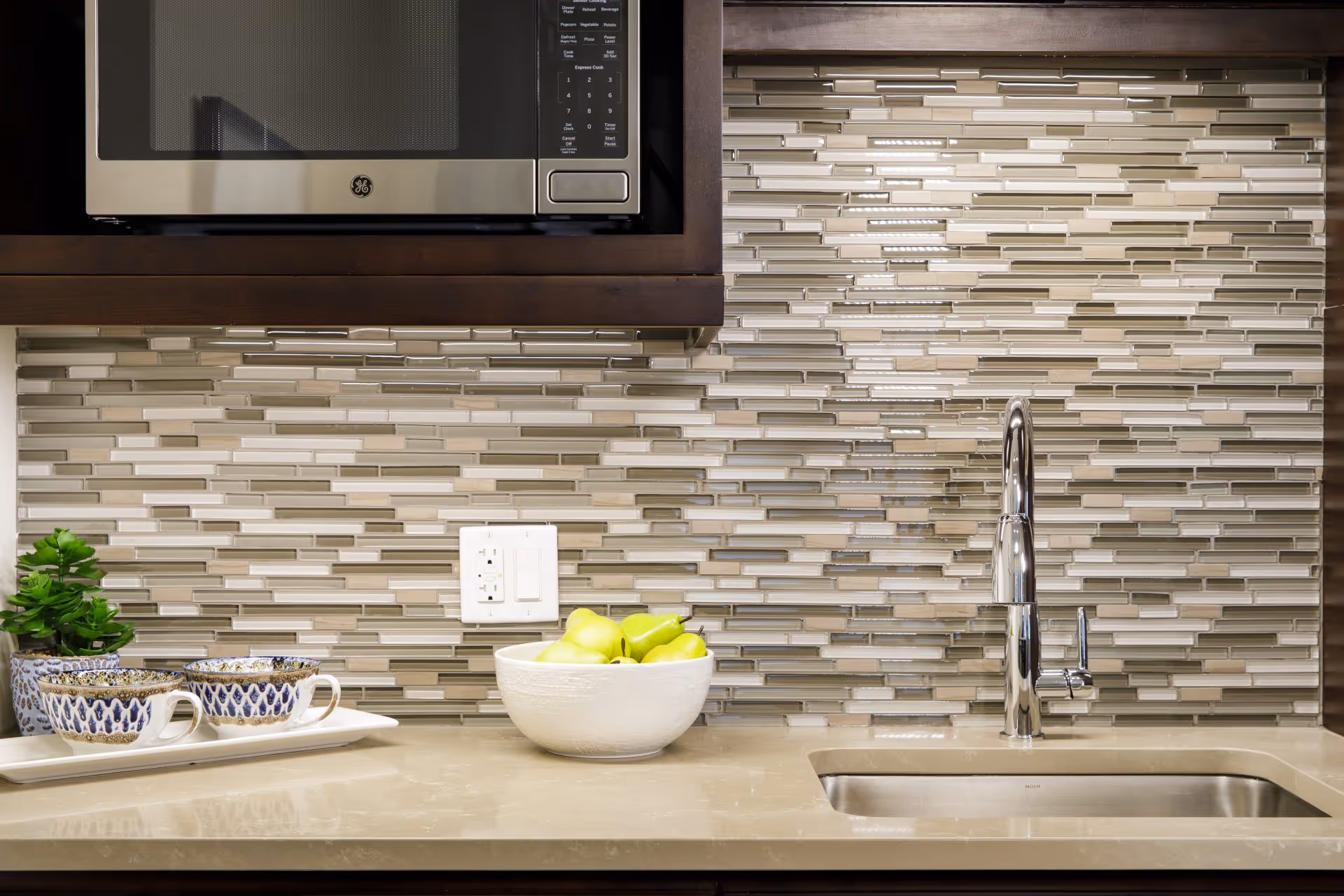 Modern kitchen countertop with a stainless steel sink and chrome faucet, a bowl of green apples, a small potted plant, and three decorative cups on a tray. Above the countertop is a stainless steel microwave and a backsplash with horizontal rectangular tiles in shades of beige, gray, and white.