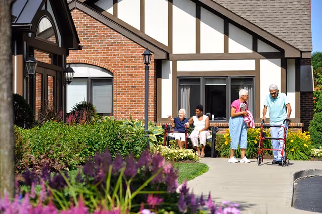 Elderly residents outside the Loudonville Assisted Living Residence—two seated on a bench and two using walkers on a garden path in front of the building.