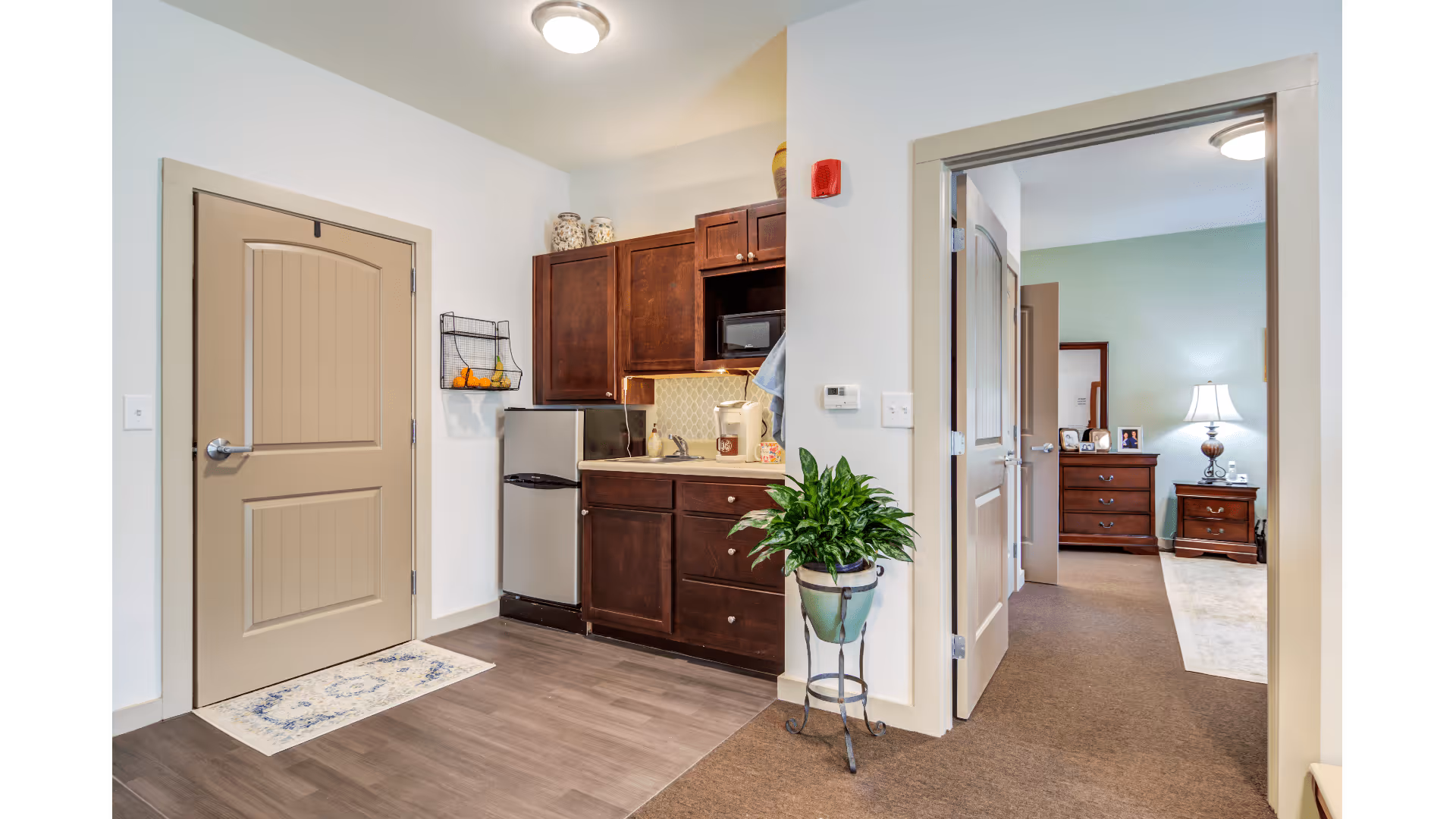Interior view of a senior living facility unit showing a small kitchenette with dark wood cabinets, a mini refrigerator, microwave, and sink. To the right, an open doorway leads to a bedroom furnished with a dresser, nightstand, lamp, and a mirror. A green potted plant is placed on a stand near the kitchenette area.