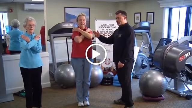 Two elderly women and a male instructor in a fitness room performing stretching exercises with their arms crossed over their chests. The room contains exercise equipment including treadmills and large exercise balls, with a wellness program poster visible in the background.
