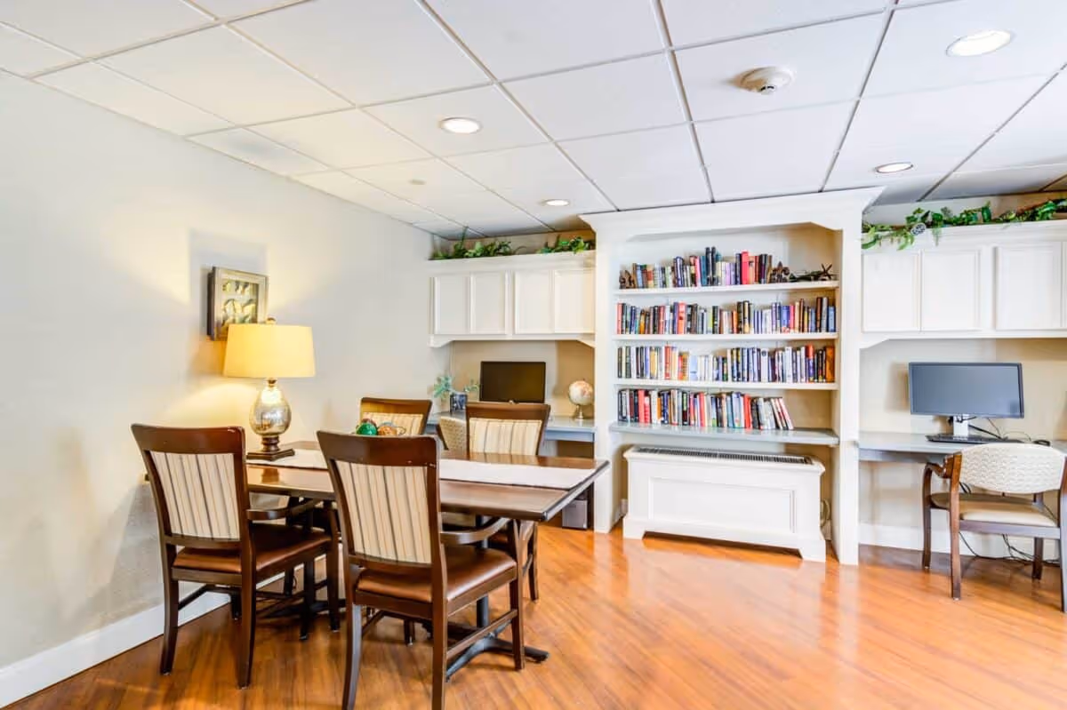 A bright room with a wooden table and four chairs in the foreground, a table lamp on the left side, and a built-in bookshelf filled with books in the background. There are two computer desks with chairs and monitors on either side of the bookshelf. The room has wooden flooring and a white ceiling with recessed lighting.