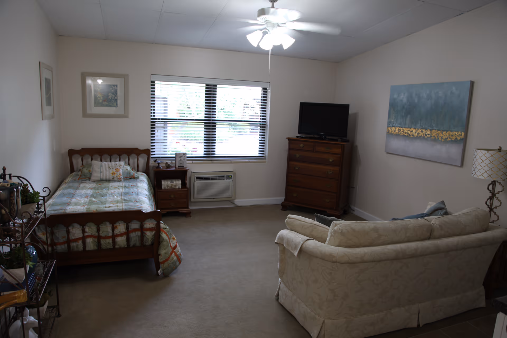 A cozy room with a single wooden bed covered with a patterned quilt, a wooden nightstand with items on it, a wooden dresser with a TV on top, a beige upholstered sofa, a floor lamp, and a ceiling fan with lights. The room has a large window with blinds and light-colored walls adorned with framed artwork.