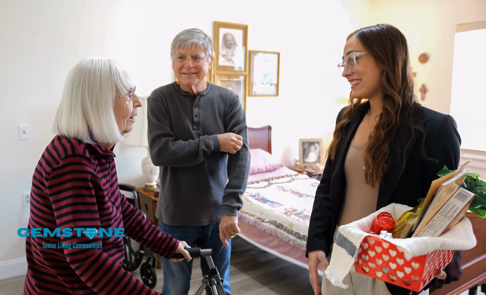 An elderly woman using a walker and an elderly man standing in a bedroom, smiling and talking with a younger woman holding a basket filled with groceries and household items. The bedroom has a bed with a floral quilt, framed pictures on the wall, and a window letting in natural light.