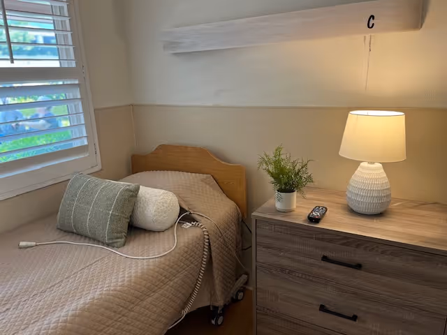 A small bedroom with a single bed covered in a beige quilt and two pillows, one rectangular and one cylindrical. Next to the bed is a wooden dresser with three drawers, a white textured lamp, a small potted plant, and a TV remote on top. A window with white blinds is on the left wall, and a white wooden beam with the letter 'C' is mounted on the wall above the dresser.