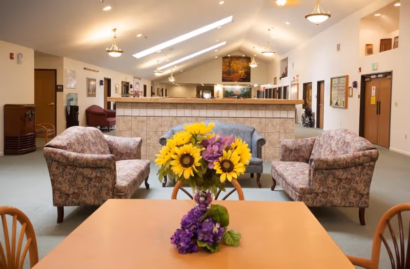 A communal lounge with a table topped by a vase of sunflowers and purple flowers, upholstered sofas, a tiled low wall, and a high skylit ceiling with chandeliers.