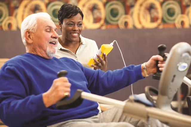 An elderly man in a blue sweater is exercising on a stationary rowing machine while a smiling woman in a beige shirt holds a yellow device and watches him.
