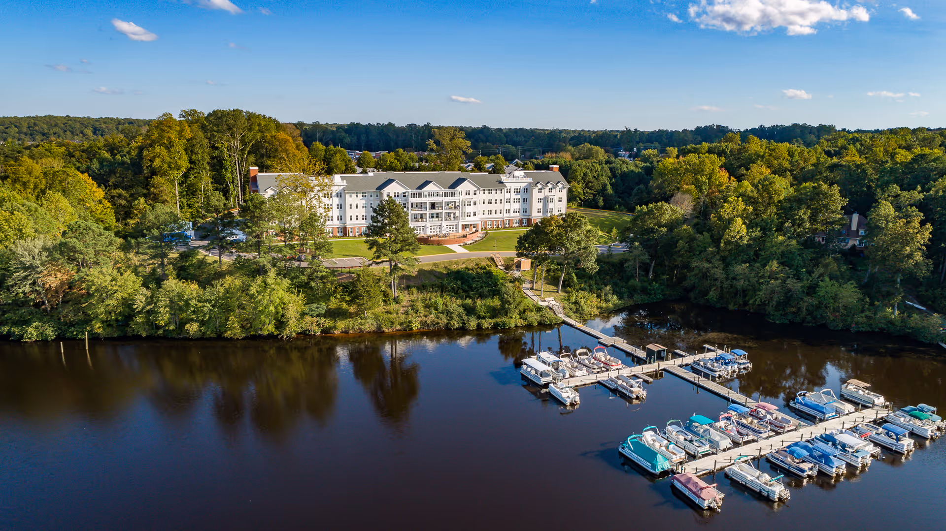 A large senior living facility building surrounded by trees, located near a calm body of water with multiple docks and boats. The sky is clear with a few clouds.
