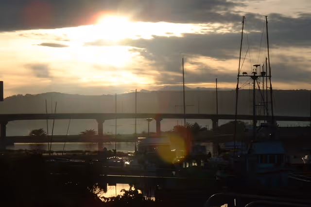 Sunset over a marina with boats and masts in the foreground and a bridge silhouetted against the sky.