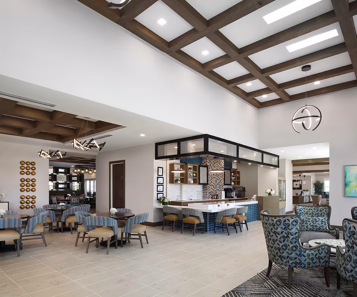 Spacious senior living communal dining and lounge area with tables, chairs, a central service counter and decorative ceiling beams.