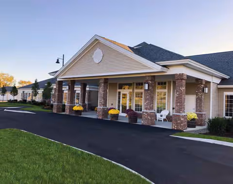 Exterior view of Woodlands Senior Living of Farmington showing a single-story building with a covered entrance supported by brick columns, a paved driveway, and some landscaping with grass and small trees.
