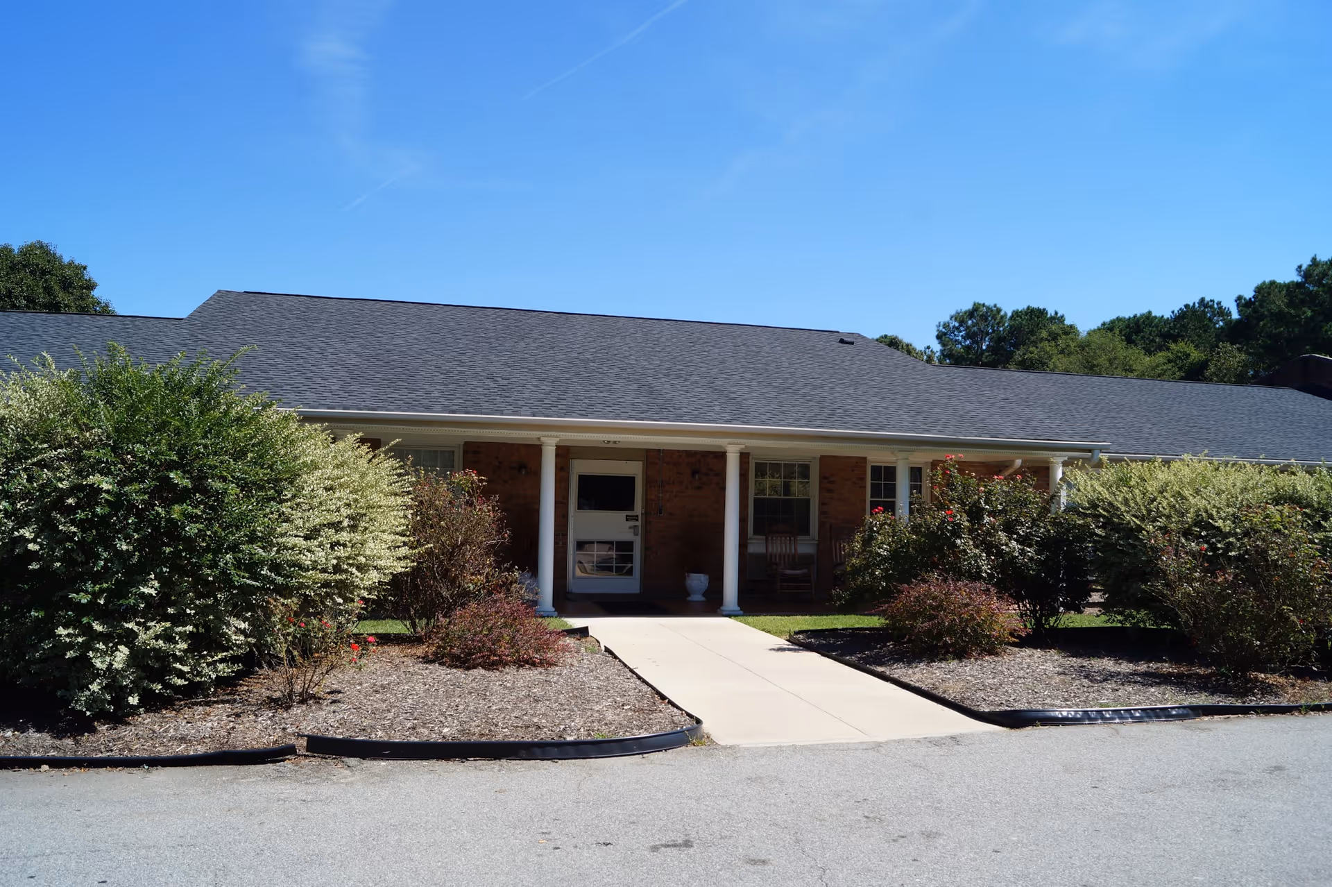 Single-story brick building entrance with a covered porch and columns, a walkway and landscaped shrubs under a clear blue sky.