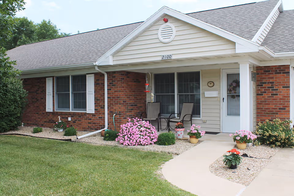 Exterior view of a single-story brick and siding building with a small covered porch. The porch has two chairs and a small table, surrounded by flower pots with pink and red flowers. The building number 2100 is displayed above the porch. There is a well-maintained lawn and landscaping with bushes and flowers along the walkway leading to the porch.