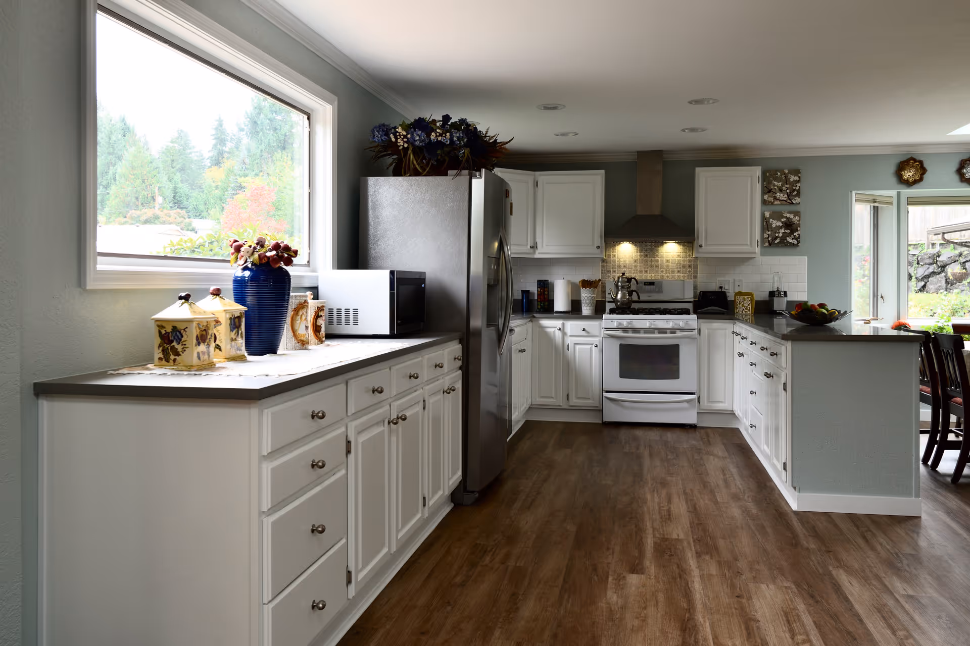A modern kitchen with white cabinets, stainless steel refrigerator, white stove with range hood, microwave, and wooden flooring. There is a large window on the left side letting in natural light, and decorative items including a blue vase and canisters on the countertop. The kitchen opens into a dining area with a table and chairs visible on the right.