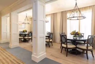 A bright dining area with round tables and chairs, each table decorated with a floral centerpiece. The room features large windows with sheer curtains allowing natural light to fill the space, and elegant chandeliers hanging from the ceiling. White paneled columns separate the dining sections.