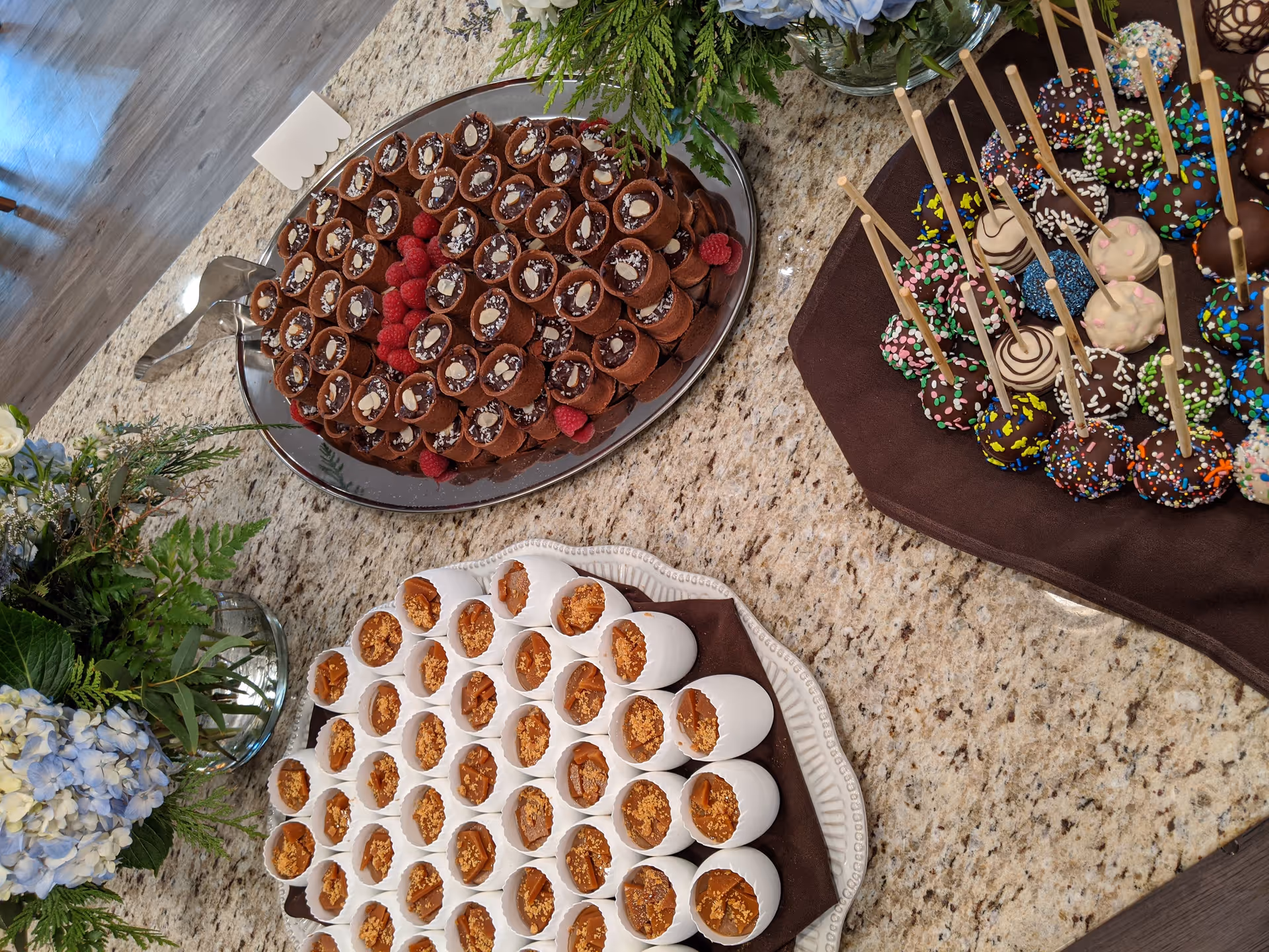 A granite countertop with three platters of assorted desserts including chocolate mousse cups garnished with raspberries, caramel-filled cups topped with crushed nuts, and colorful cake pops with sprinkles. There are also floral arrangements with green leaves and light blue flowers on the countertop.