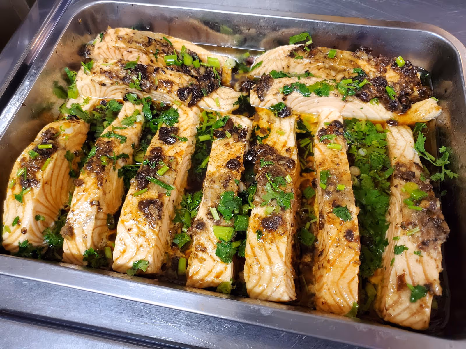 Tray of cooked salmon fillets garnished with chopped green herbs and sauce, arranged neatly in a stainless steel serving tray.