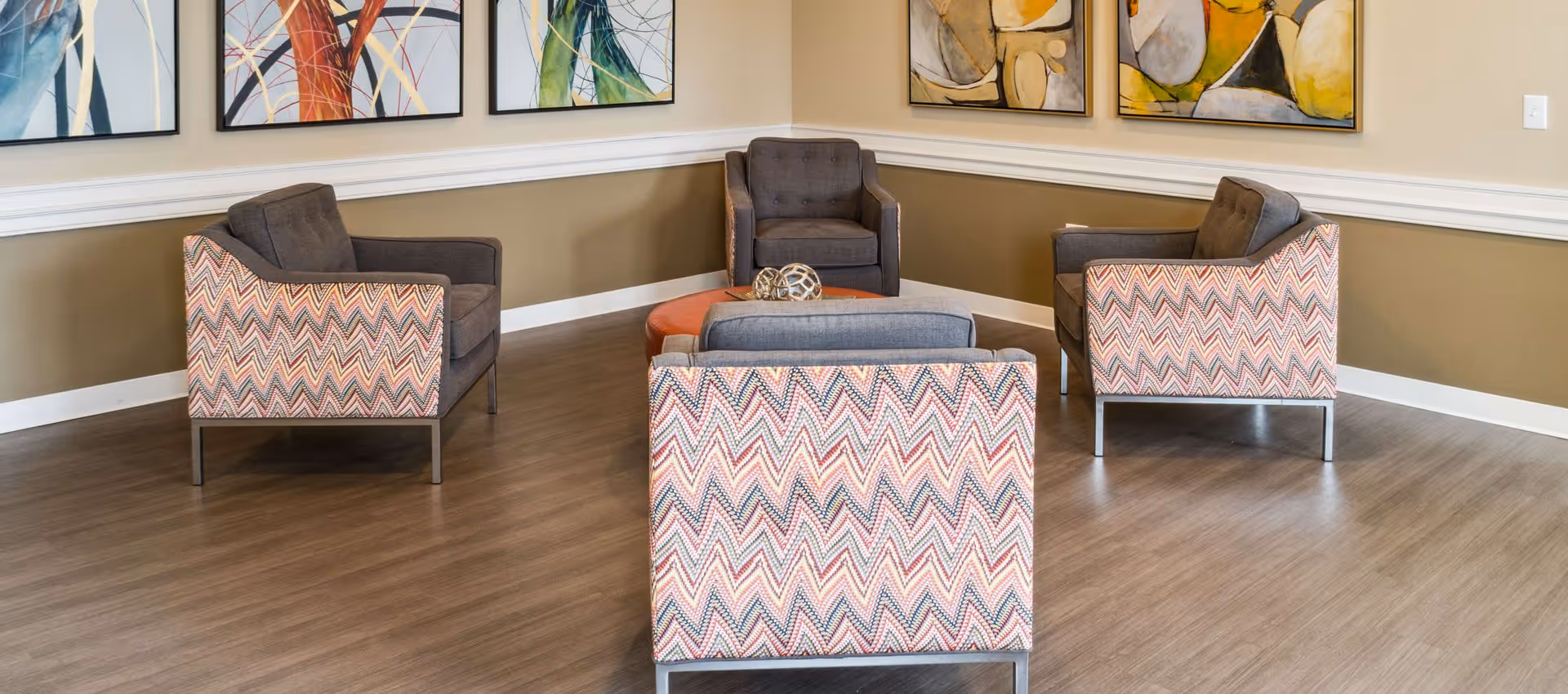 A cozy seating area with four armchairs arranged around a round ottoman in the center. The armchairs have a patterned fabric on the sides and back, with solid gray cushions. The walls are beige with white trim and decorated with colorful abstract paintings.