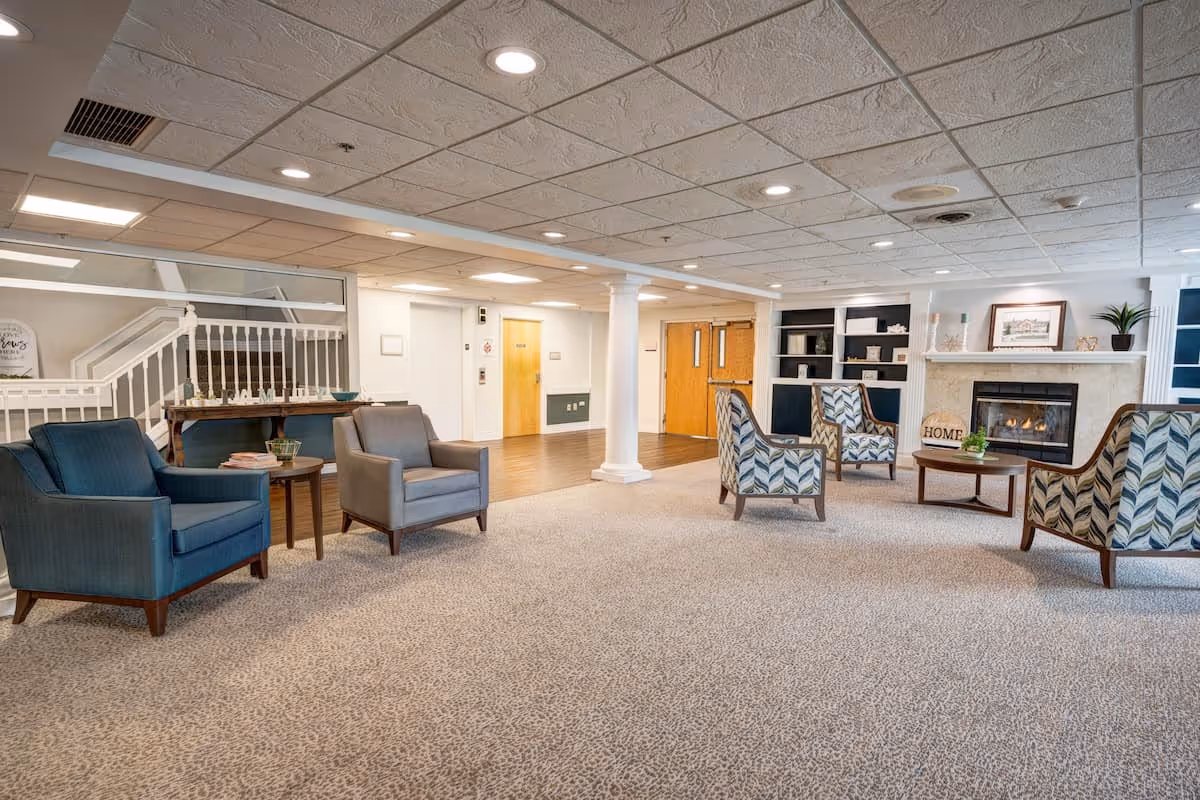 A spacious and well-lit living room area in a senior living facility with multiple armchairs arranged around a coffee table and a fireplace. The room features carpeted flooring, a white column, built-in shelves with decorative items, and a staircase with a white railing in the background.