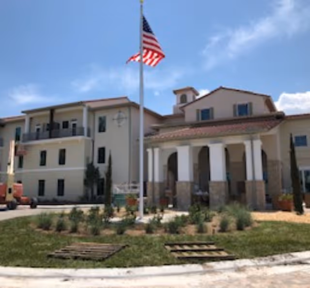 Exterior view of Tuscan Gardens Of Delray Beach senior living facility showing a multi-story building with balconies, a covered entrance with columns, an American flag on a flagpole, and landscaped greenery under a partly cloudy sky.