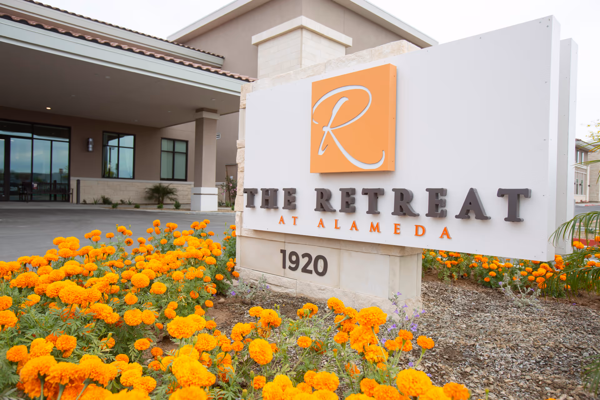 The entrance sign for The Retreat at Alameda, featuring a large white and stone base with the facility's name and address 1920, surrounded by orange flowers and a covered driveway entrance to the building in the background.