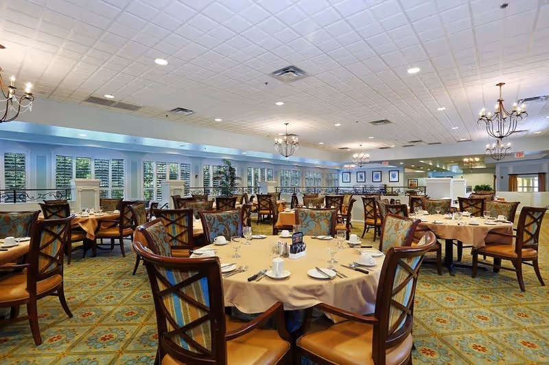A spacious dining room with multiple round tables covered with beige tablecloths, each set with white cups, plates, silverware, and glassware. The room features patterned carpet flooring, wooden chairs with cushioned seats and backs, chandeliers hanging from a white tiled ceiling, and large windows with white shutters along one wall.