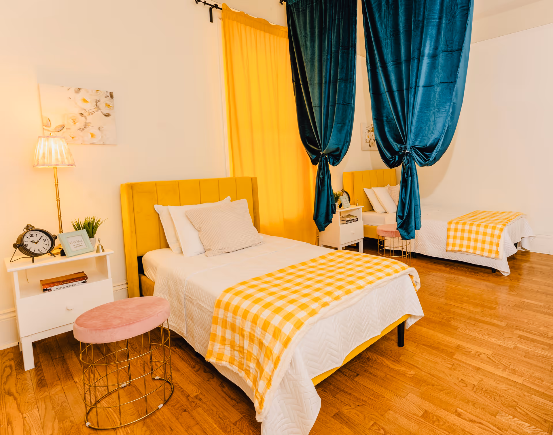 A bright bedroom with two single beds featuring yellow upholstered headboards and white bedding with yellow checkered blankets. Each bed has a white nightstand beside it, decorated with a lamp, clock, small plant, and picture frame. The room has wooden flooring, yellow curtains, and teal velvet curtains hanging from the ceiling between the beds.