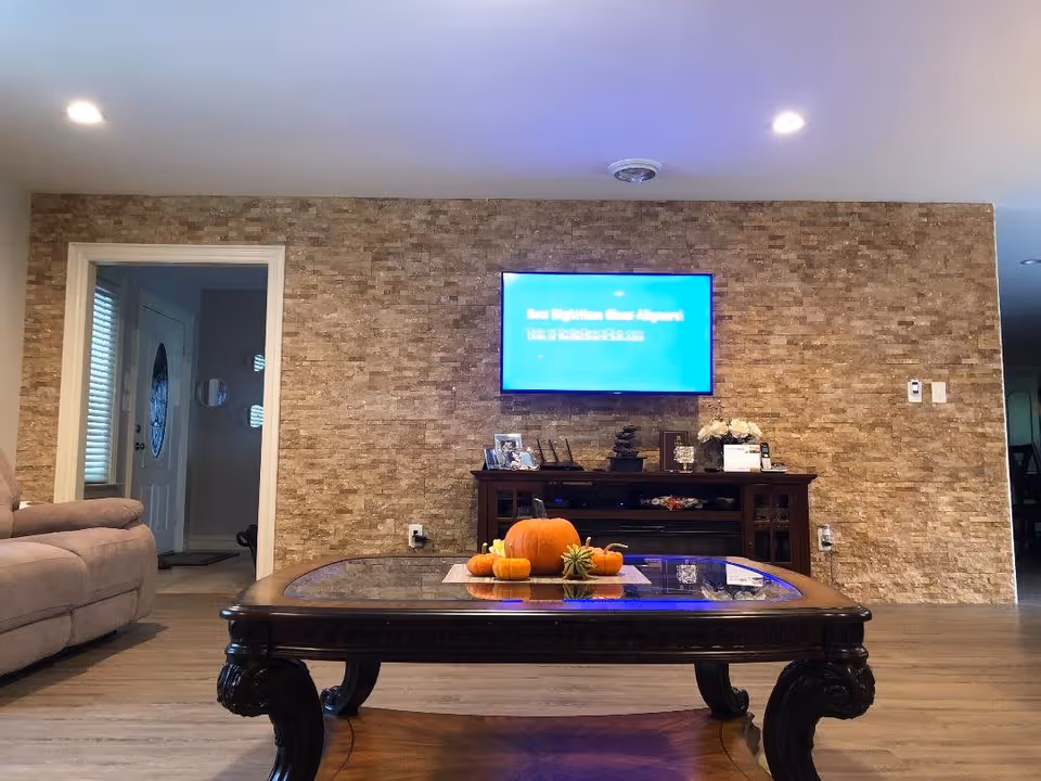 A living room with a stone-textured accent wall, a mounted flat-screen TV displaying a blue screen, a dark wooden TV stand with decorative items, and a wooden coffee table with small pumpkins and a succulent on it. A beige sofa is partially visible on the left side, and a doorway leads to another room with a white door and windows with blinds.