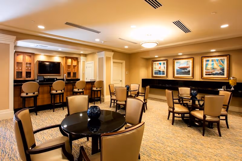 A well-lit common area with multiple round tables and beige cushioned chairs. There is a bar counter with four high chairs and a TV mounted on the wall behind it. The walls are decorated with three framed paintings of sailboats. The room has a warm, inviting atmosphere with carpeted flooring and recessed lighting.