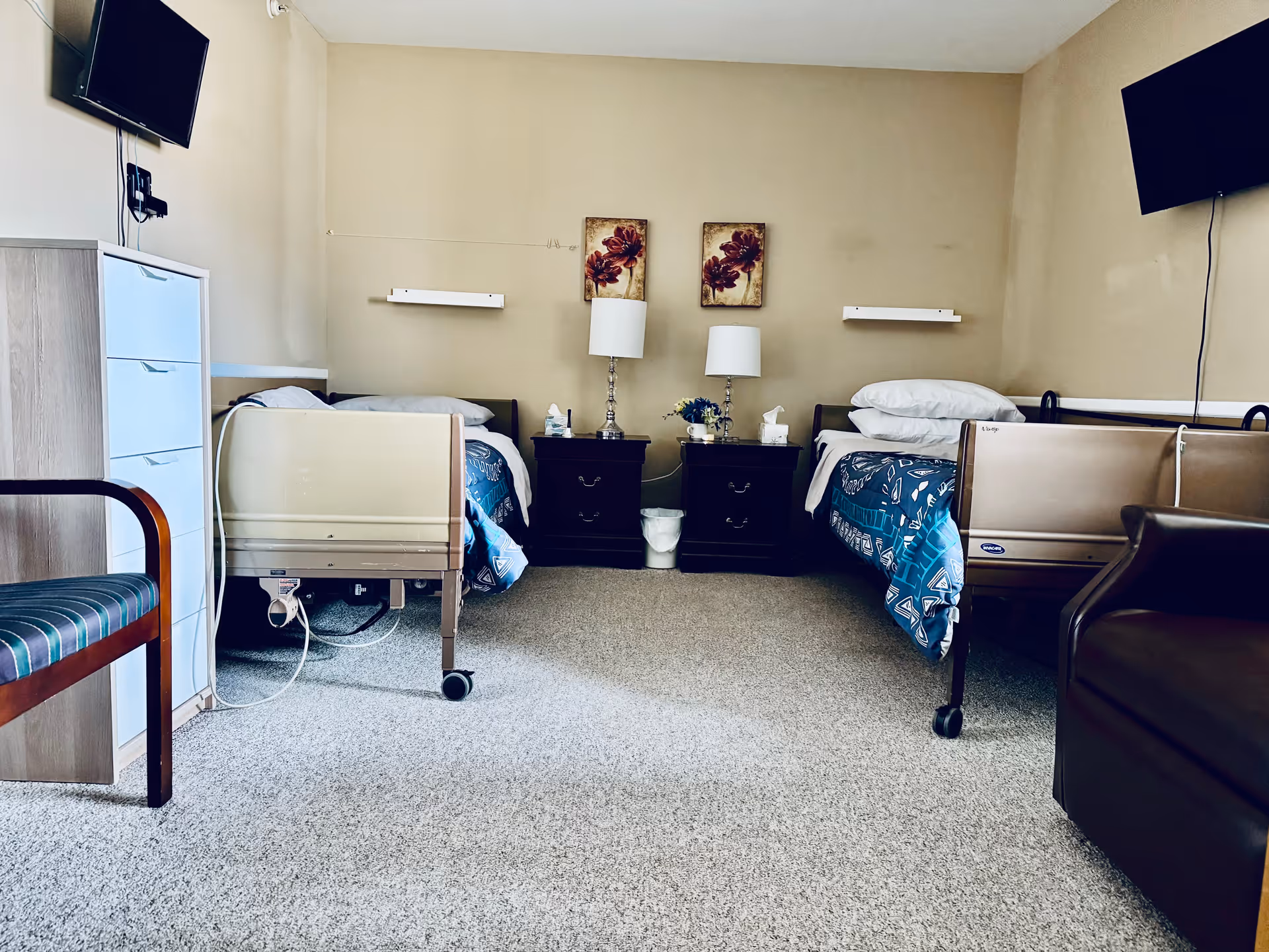 A room with two hospital-style beds separated by two dark wooden nightstands with lamps and decorative flowers. Each bed has a blue patterned blanket and white pillows. There are two wall-mounted televisions above each bed. A striped chair is visible on the left side and a brown armchair on the right side. The walls are beige and the floor is carpeted.