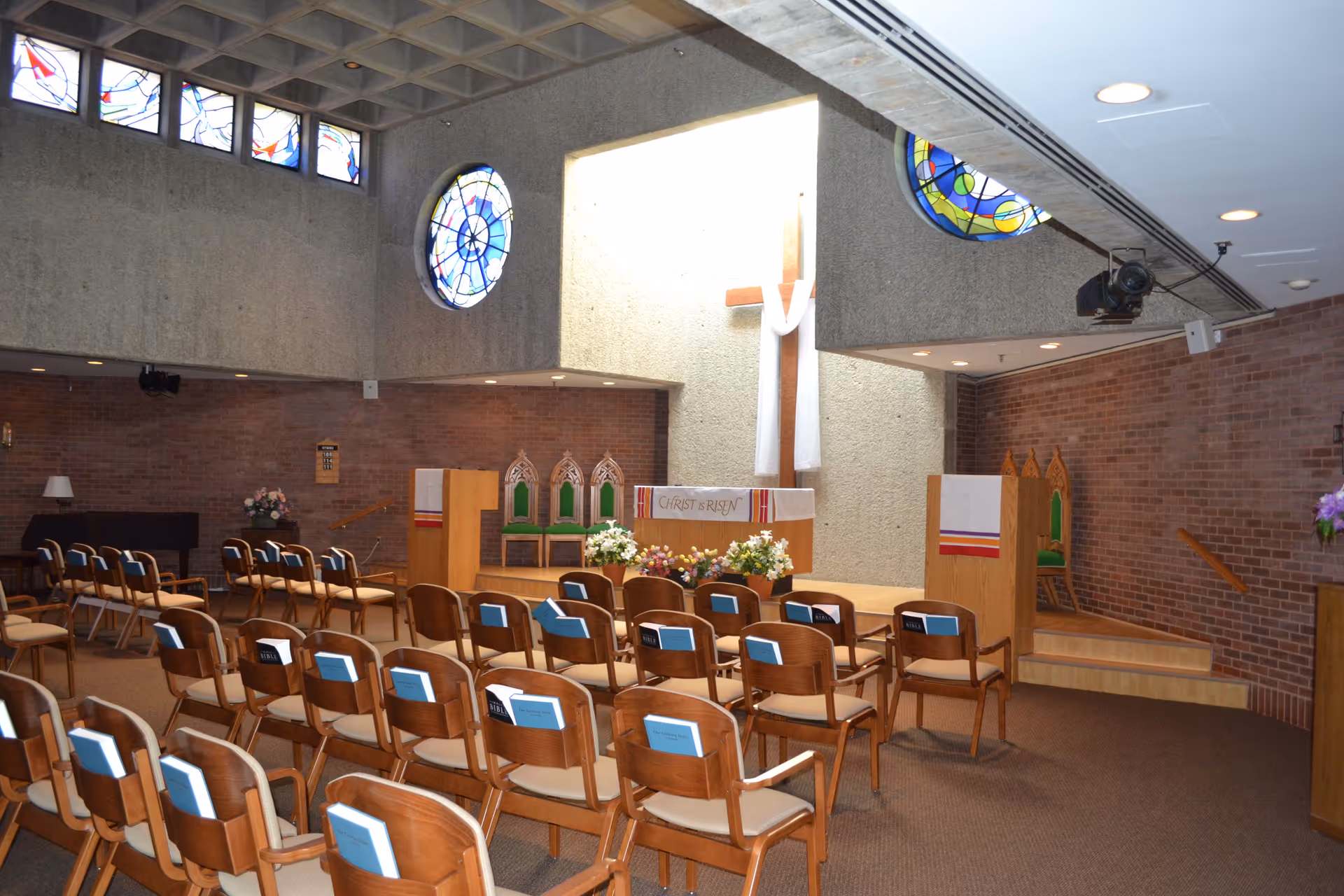 Interior view of a chapel with rows of wooden chairs facing an altar. The altar has a white cloth with the words 'CHRIST IS RISEN' and is decorated with flowers. Behind the altar is a large wooden cross draped with white fabric. The chapel has stained glass windows and brick walls.
