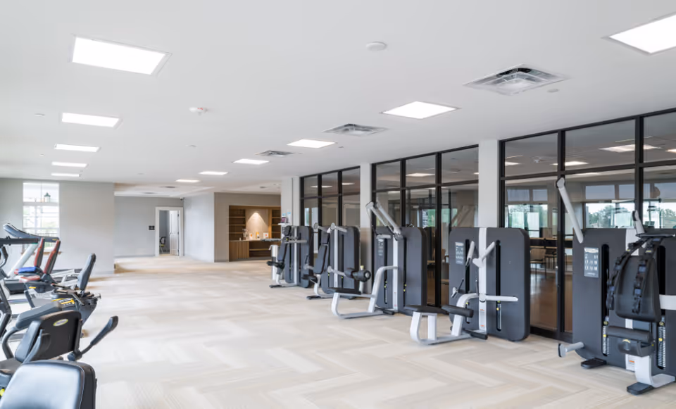A spacious and well-lit fitness room with various exercise machines including stationary bikes and strength training equipment. The room has large windows and glass walls separating it from an adjacent room with tables and chairs.