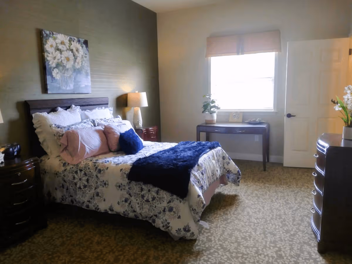 A cozy bedroom with a bed featuring floral bedding and multiple pillows, a bedside table with a lamp, a window with a blind, a small table with a plant and picture frame, and a dresser with a vase of flowers.