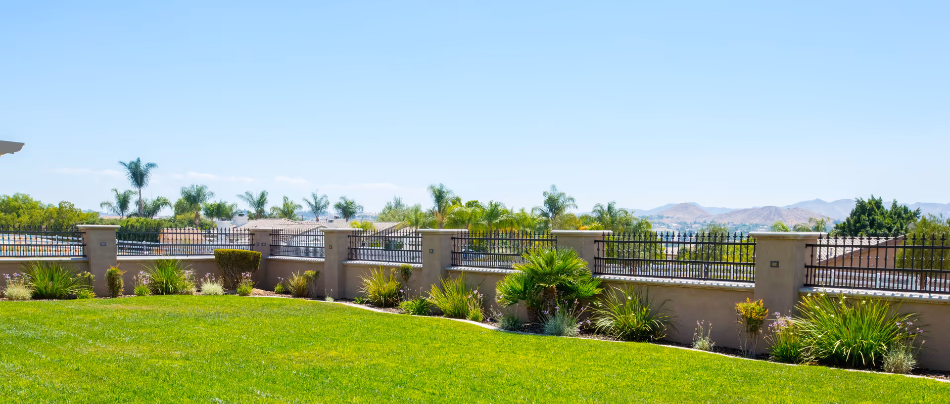 Well-maintained green lawn and landscaped garden with a low stucco wall and iron fence, palm trees and distant hills under a clear blue sky.