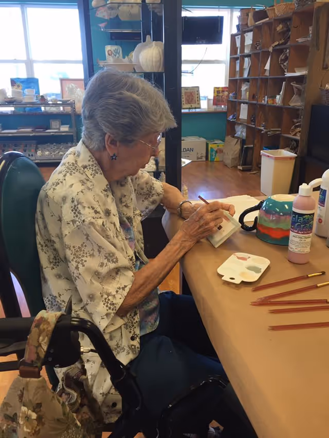 An elderly woman with gray hair and glasses is sitting at a table in a craft room, painting a small object. She is wearing a floral patterned shirt and has a walker with a floral bag attached beside her. The table has paintbrushes, paint bottles, and a paint palette. Shelves with various craft supplies and decorations are visible in the background.