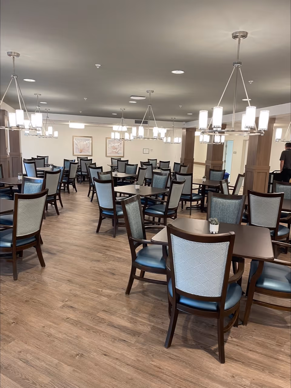 A spacious dining room with multiple dark wood tables and chairs with blue cushioned seats and patterned backs. The room has wooden flooring, modern hanging light fixtures, and light-colored walls adorned with framed artwork. A person is seen in the background near a doorway.