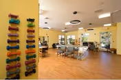 A spacious rehabilitation and healthcare center room with wooden flooring and light yellow walls. On the left wall, there is a vertical rack holding colorful dumbbells. The room contains several tables and chairs arranged for therapy or activities, with large windows allowing natural light to enter. Ceiling lights and fans are visible, and the overall atmosphere is bright and welcoming.