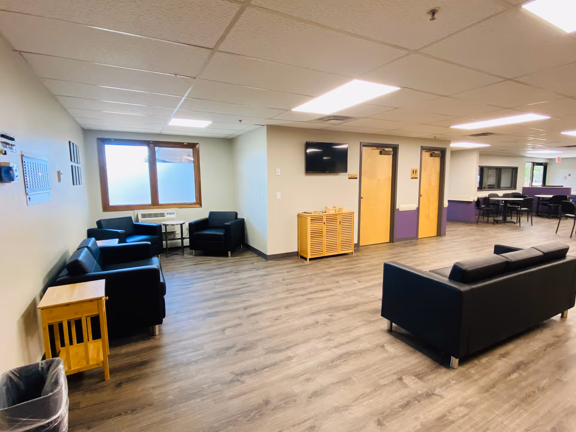 A spacious common area with wood flooring and a drop ceiling with fluorescent lights. The room features black leather sofas and armchairs arranged around small tables. There is a wooden cabinet under a wall-mounted TV, and two wooden doors with restroom signs. In the background, there are tables and chairs near windows.